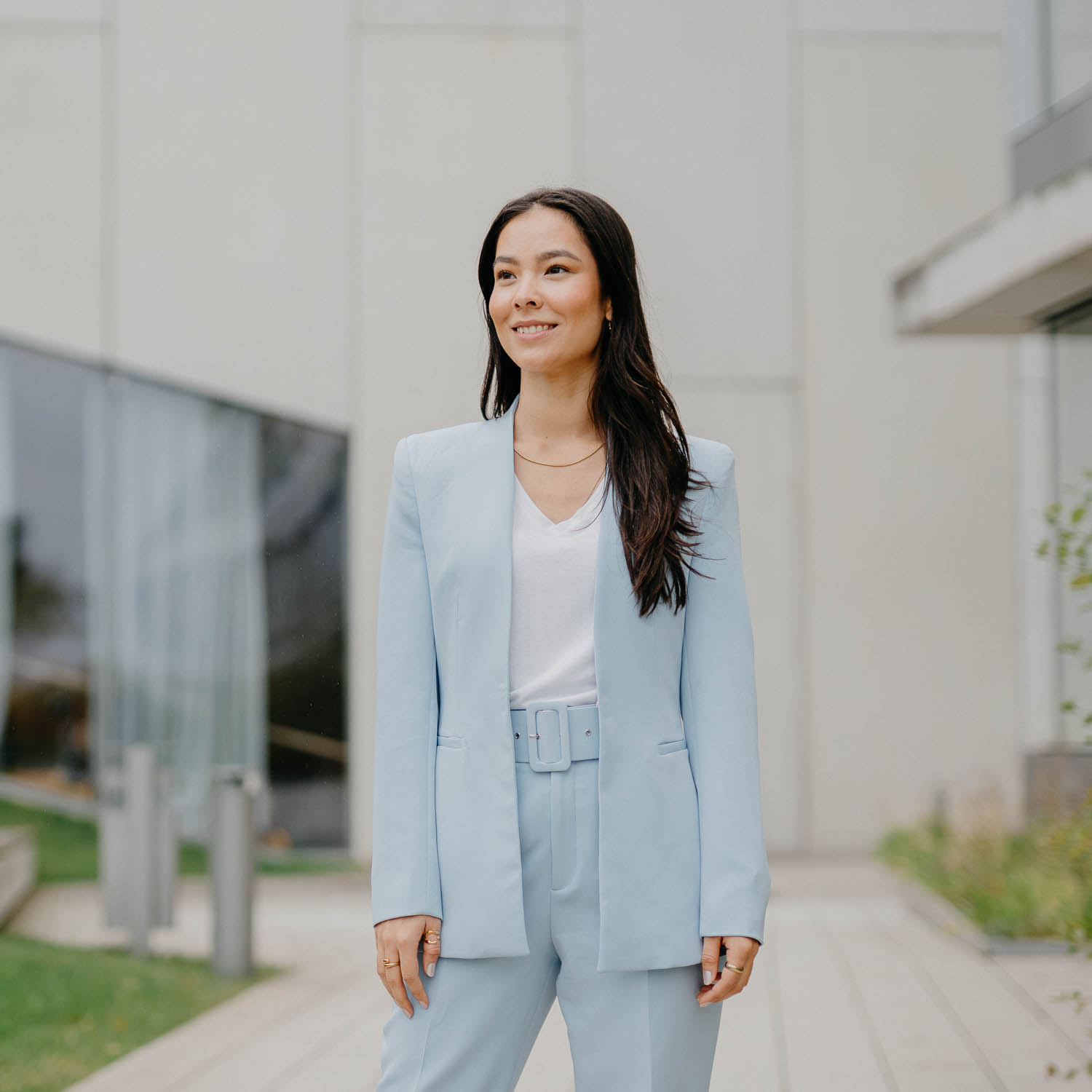 Frau im hellblauen Hosenanzug lächelt selbstbewusst vor modernem Bürogebäude — stehendes Businessportrait.
