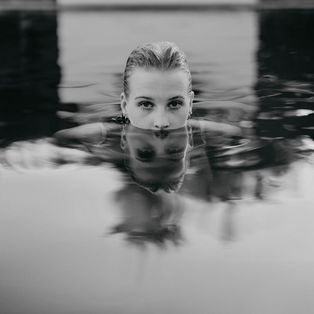 Porträt einer Frau, die bis zur Nase im Wasser eines Pools eingetaucht ist, mit klarem Blick in die Kamera und ihrer Spiegelung auf der Wasseroberfläche, in Schwarz-Weiß aufgenommen.