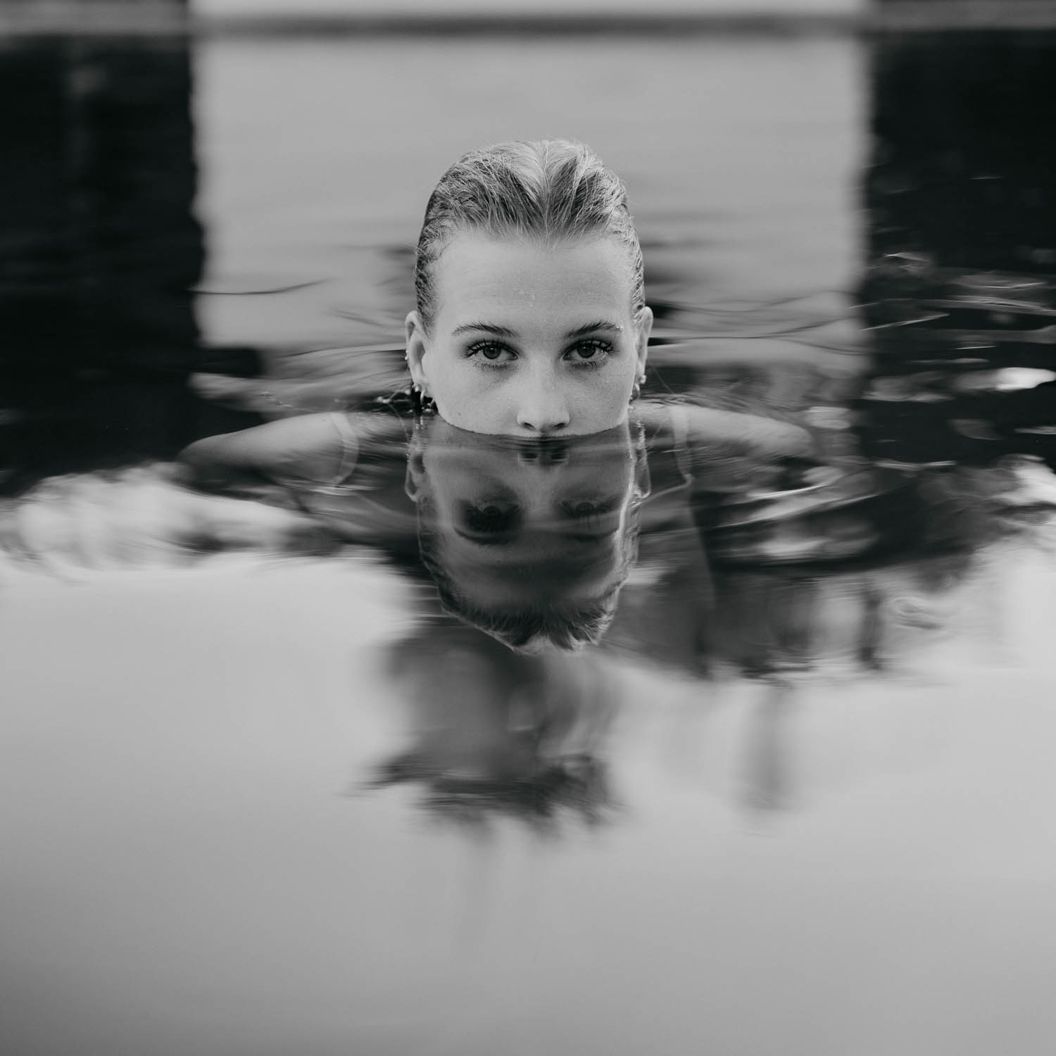 Porträt einer Frau, die bis zur Nase im Wasser eines Pools eingetaucht ist, mit klarem Blick in die Kamera und ihrer Spiegelung auf der Wasseroberfläche, in Schwarz-Weiß aufgenommen.