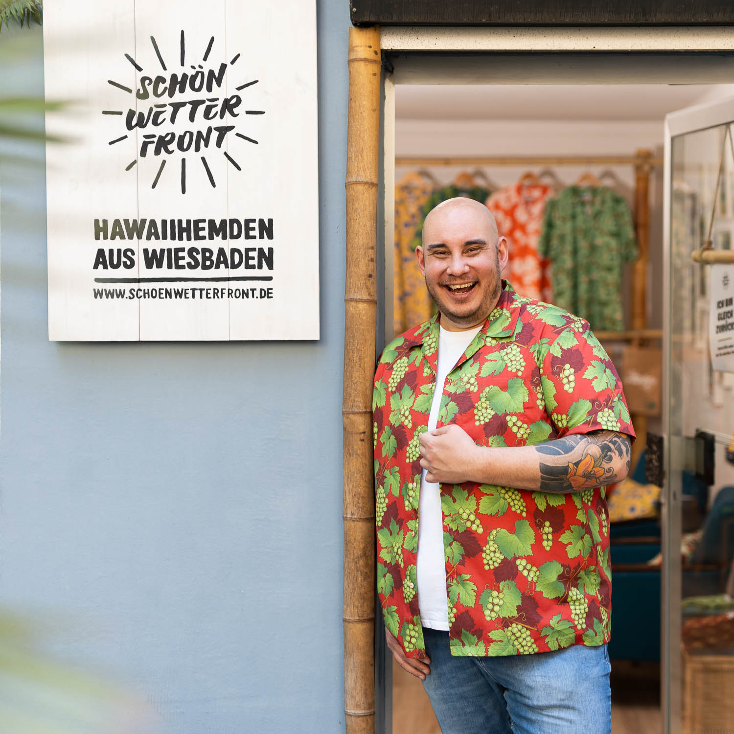 Christian Jakob, Inhaber vom Modelabel Schönwetterfront mit gemustertem Hawaiihemd steht lächelnd vor seinem Laden und zeigt auf ein Holzschild mit der Aufschrift „Schönwetterfront – Hawaiihemden aus Wiesbaden“.