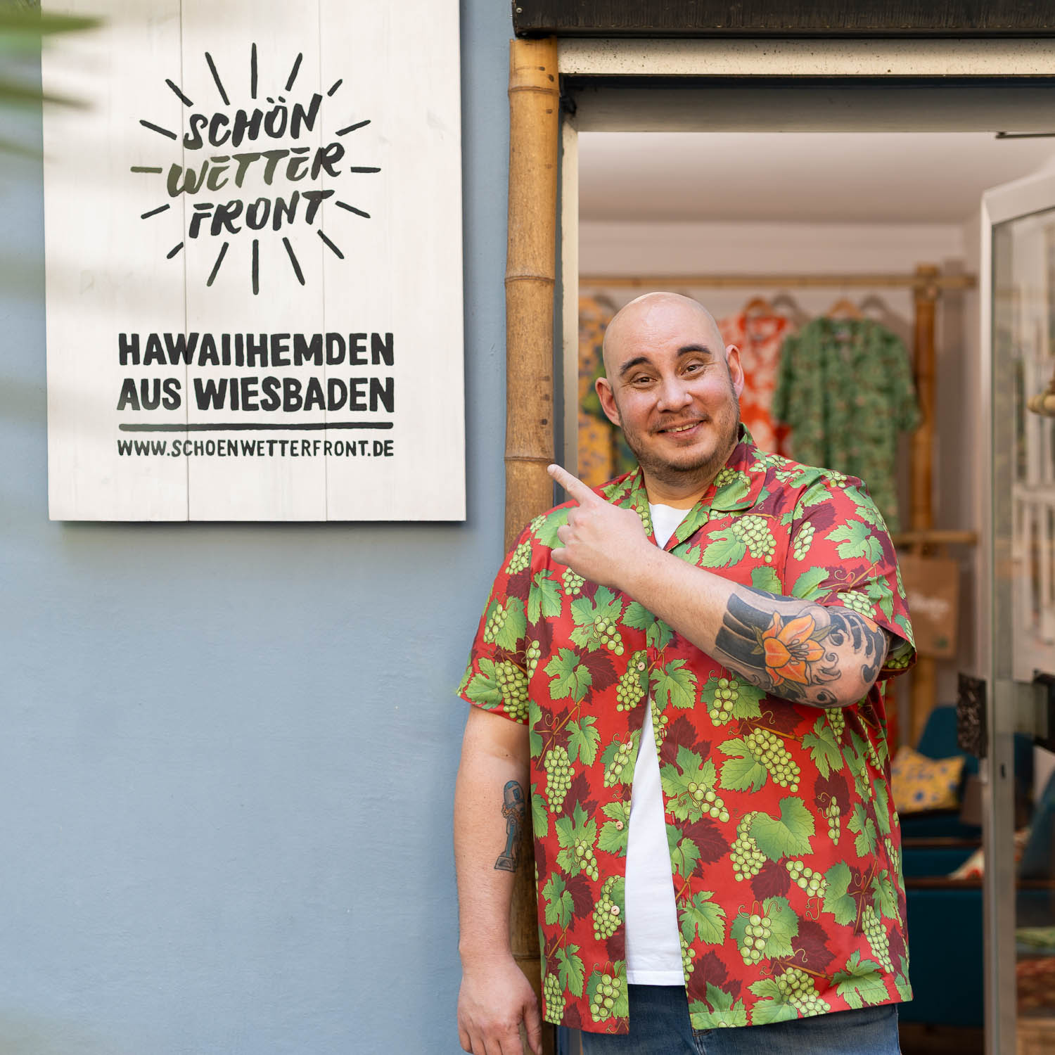 Christian Jakob, Inhaber vom Modelabel Schönwetterfront mit gemustertem Hawaiihemd steht lächelnd vor seinem Laden und zeigt auf ein Holzschild mit der Aufschrift „Schönwetterfront – Hawaiihemden aus Wiesbaden“.