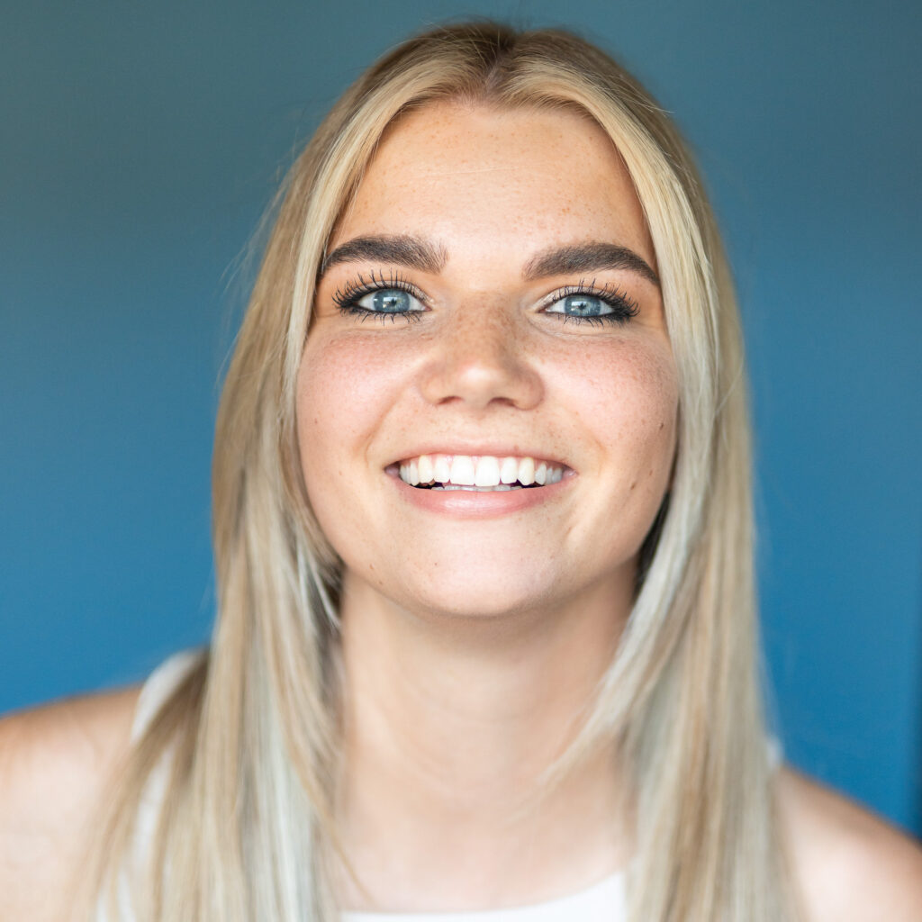 Porträt einer jungen Frau mit blonden Haaren und blauen Augen, die herzlich in die Kamera lächelt.