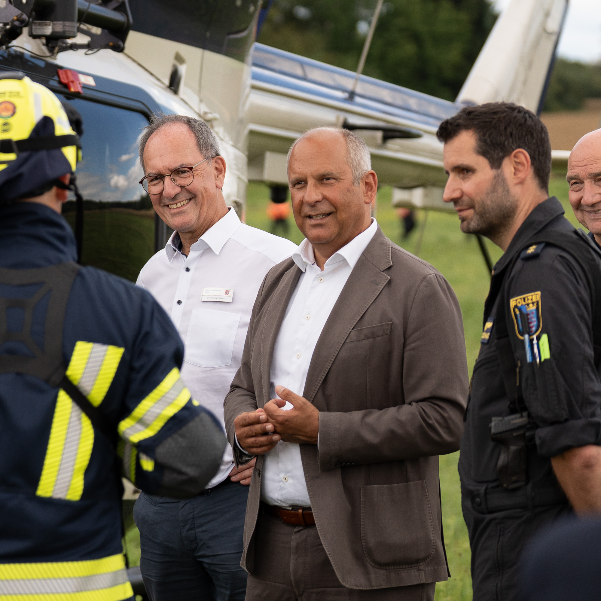 Mehrere Personen, darunter Vertreter aus Politik, Polizei und Feuerwehr, im Gespräch vor einem Hubschrauber auf einer Wiese.