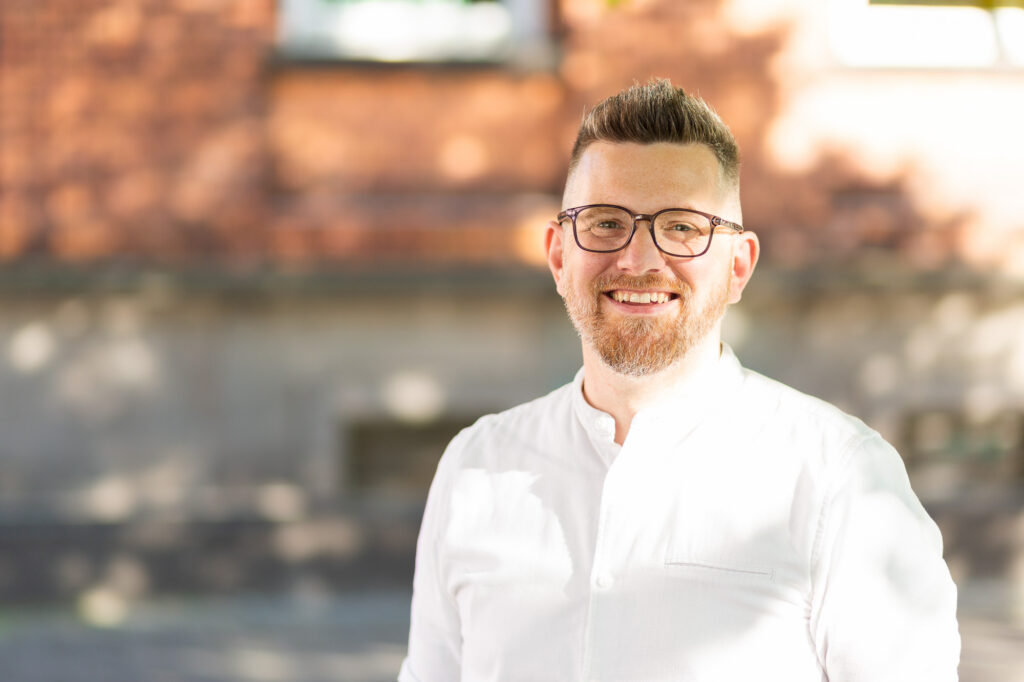 Porträt eines Mannes mit Brille und tätowiertem Unterarm, der in weißem Hemd und Jeans draußen vor einem Backsteingebäude steht und freundlich lächelt.