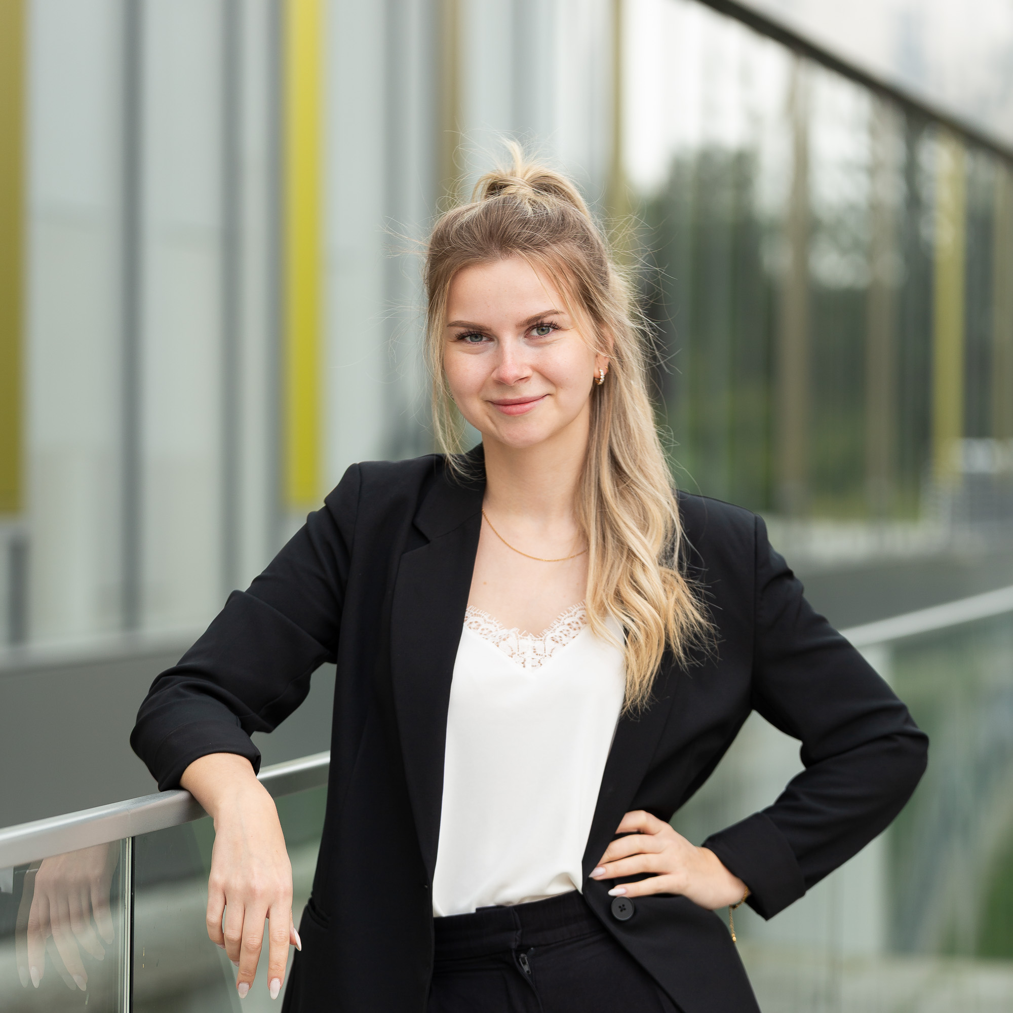 Bewerbungsfoto einer jungen Frau in Businesskleidung, die selbstbewusst an einem Glasgeländer steht und in die Kamera lächelt.