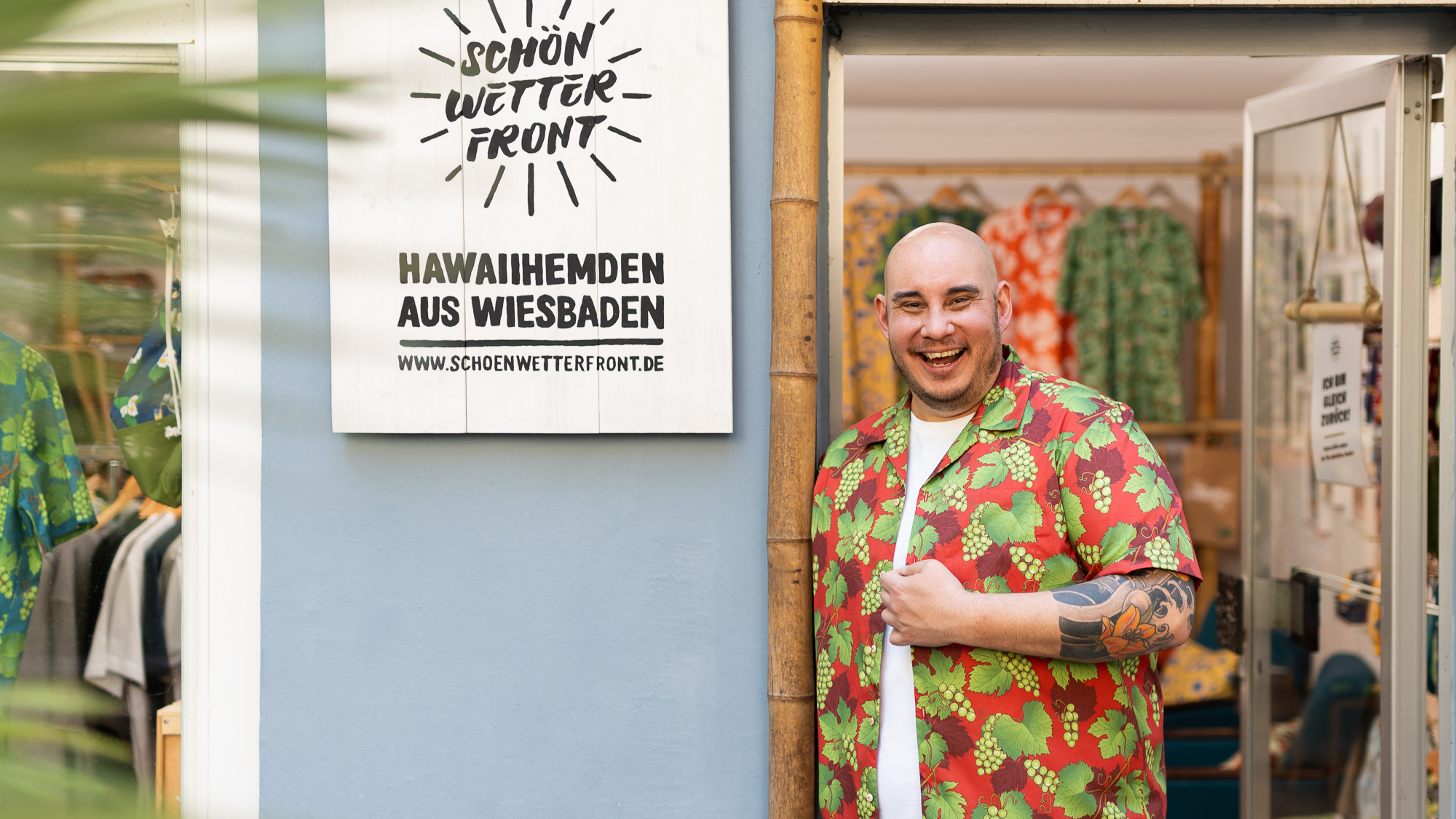Christian Jakob, Inhaber vom Modelabel Schönwetterfront mit gemustertem Hawaiihemd steht lächelnd vor seinem Laden und zeigt auf ein Holzschild mit der Aufschrift „Schönwetterfront – Hawaiihemden aus Wiesbaden“.