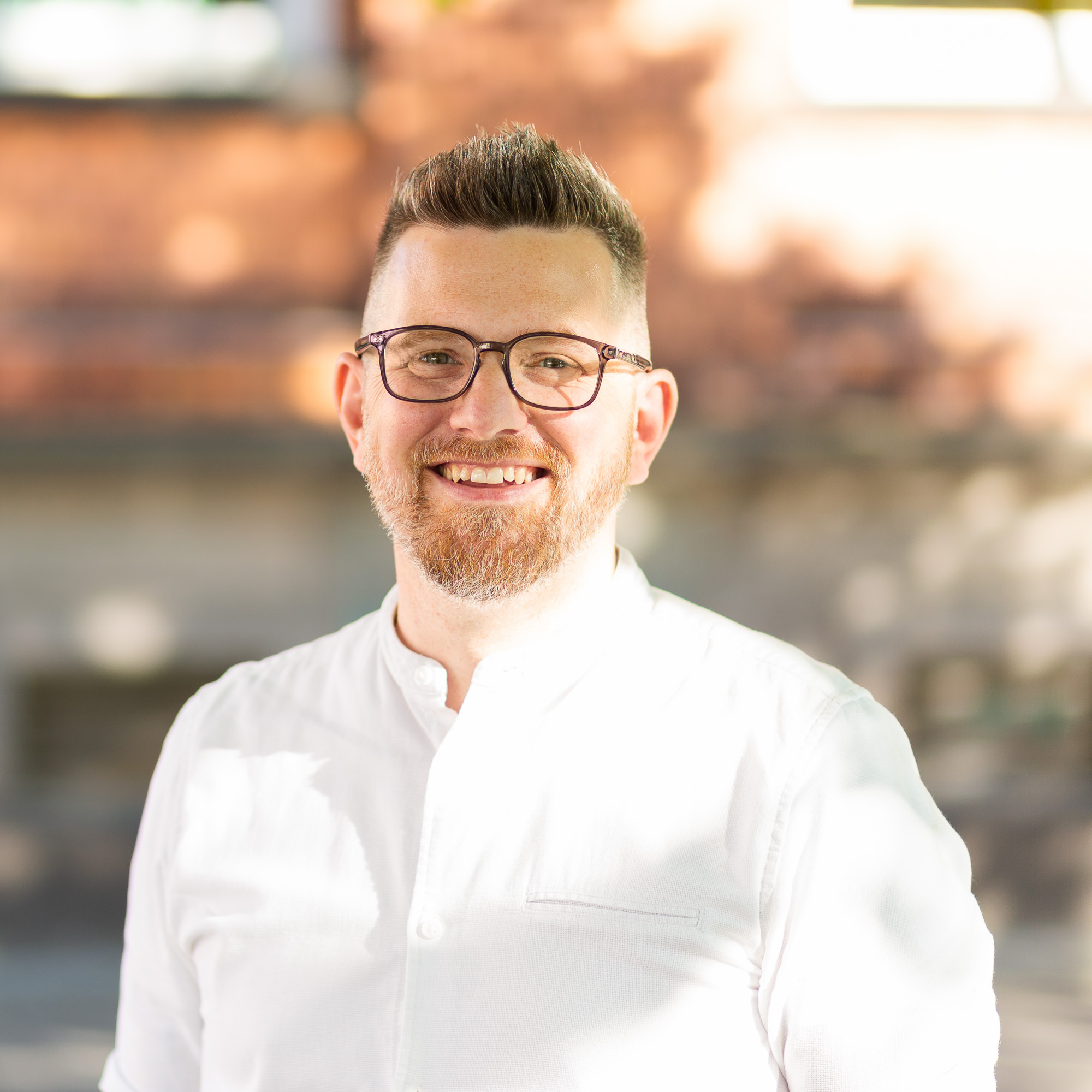 Porträt eines Mannes mit Brille und tätowiertem Unterarm, der in weißem Hemd und Jeans draußen vor einem Backsteingebäude steht und freundlich lächelt.