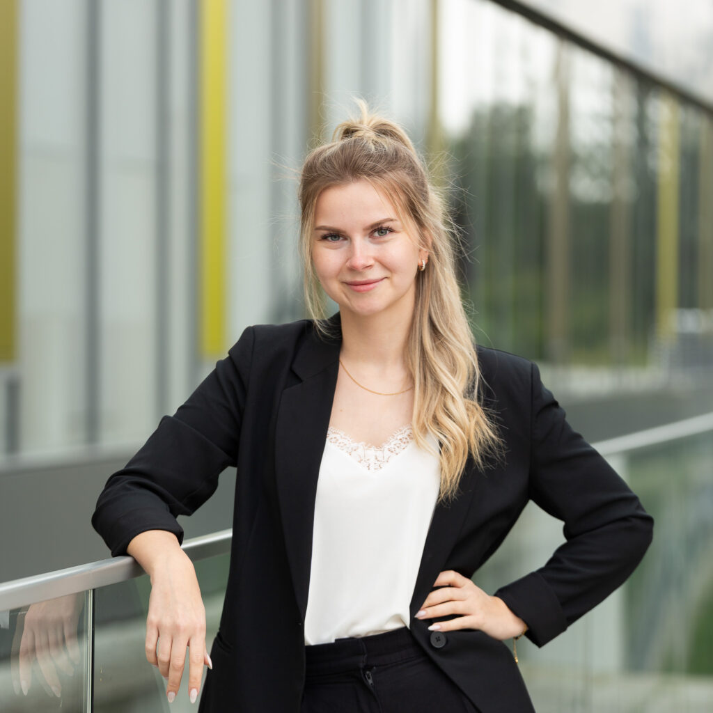 Bewerbungsfoto einer jungen Frau in Businesskleidung, die selbstbewusst an einem Glasgeländer steht und in die Kamera lächelt.