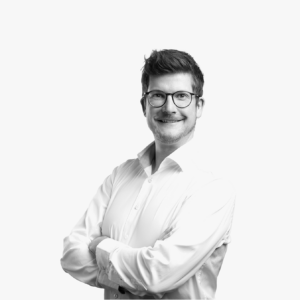 Porträt von Marcel Cornelius lächelnden mit Brille in einem weißen Hemd vor hellem Hintergrund.