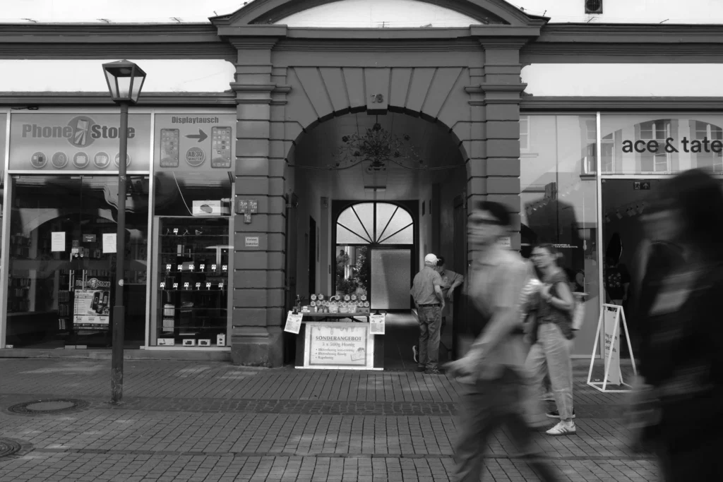 Schwarz-Weiß-Aufnahme einer historischen Häuserfassade mit Torbogen und vorbeigehenden, bewegungsunscharfen Passanten.
